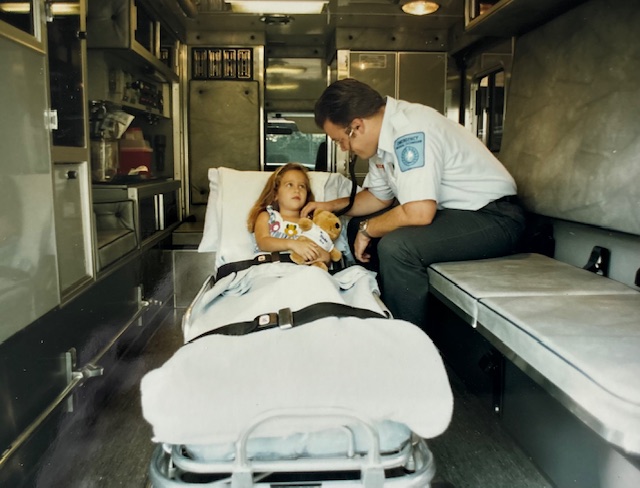 A Medix paramedic sits with a young girl patient in the back of an ambulance; she holds a stuffed teddy bear and looks up at him calmly